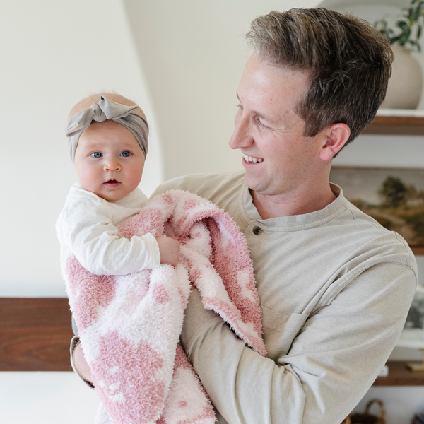 A father holds his baby girl who is holding a double-layer Bamboni® baby blanket. The plush, breathable blanket features a two-tone light pink and pink floral design, offering ultimate softness and warmth. Made from Saranoni’s signature stretchy, cloud-like Bamboni® fabric, this luxury baby blanket provides comfort for naps, tummy time, and on-the-go snuggles. The soft blanket is a receiving blanket and Saranoni blanket. 