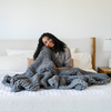 A woman smiles and sits on a bed with a soft, gray Saranoni chunky knit throw blanket. The large knit Saranoni blanket, is made from 100% poly microfiber and drapes beautifully. This luxury throw blanket is both trendy and durable, making it the perfect addition to any space. Ideal for home decor enthusiasts and those seeking a cuddly, soft blanket, this chunky knit blanket combines style and comfort. Keywords: chunky knit throw, soft blanket, knit blanket, large knit blanket, luxury throw blanket.