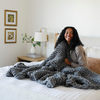 A woman smiles and sits with a soft, gray Saranoni chunky knit throw blanket. The large knit Saranoni blanket, is made from 100% poly microfiber and drapes beautifully. This luxury throw blanket is both trendy and durable, making it the perfect addition to any space. Ideal for home decor enthusiasts and those seeking a cuddly, soft blanket, this chunky knit blanket combines style and comfort. Keywords: chunky knit throw, soft blanket, knit blanket, large knit blanket, luxury throw blanket.