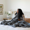 A woman sits with a soft, gray Saranoni chunky knit throw blanket. The large knit Saranoni blanket, is made from 100% poly microfiber and drapes beautifully. This luxury throw blanket is both trendy and durable, making it the perfect addition to any space. Ideal for home decor enthusiasts and those seeking a cuddly, soft blanket, this chunky knit blanket combines style and comfort. Keywords: chunky knit throw, soft blanket, knit blanket, large knit blanket, luxury throw blanket.