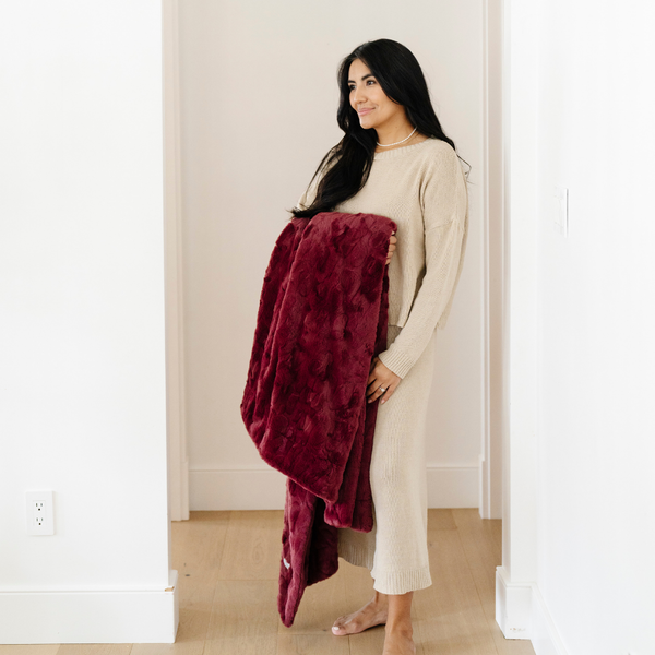 A woman smiles and holds a pomegranate luxurious patterned faux fur throw blanket that is a Saranoni blanket. This ultra-soft, plush throw blanket adds a touch of elegance and warmth to any space, perfect as a soft throw blanket or fluffy blanket for lounging. Ideal for those seeking a high-quality artificial fur blanket or luxury blanket, this versatile and stylish soft blanket enhances both comfort and decor. Shop the best blankets and throws for ultimate relaxation and cozy vibes.