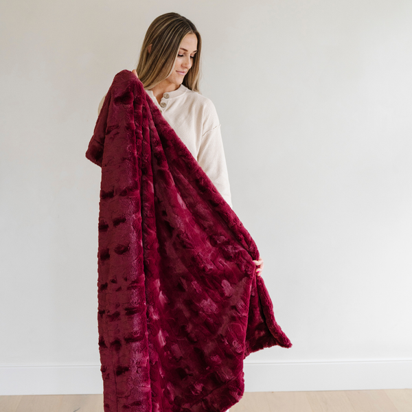 A woman holds up a pomegranate colored luxurious patterned faux fur extra large throw blanket that is a Saranoni blanket. This ultra-soft, cozy blanket adds a touch of elegance and warmth to any space, perfect as a soft oversized blanket or fluffy blanket for lounging. Ideal for those seeking a high-quality artificial fur blanket or luxury blanket, this versatile and stylish soft blanket enhances both comfort and decor. Shop the best blankets and throws for ultimate relaxation and cozy vibes.