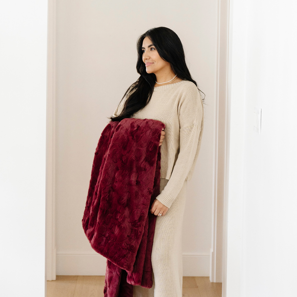 A woman smiles and holds a pomegranate colored luxurious patterned faux fur extra large throw blanket that is a Saranoni blanket. This ultra-soft, cozy blanket adds a touch of elegance and warmth to any space, perfect as a soft oversized blanket or fluffy blanket for lounging. Ideal for those seeking a high-quality artificial fur blanket or luxury blanket, this versatile and stylish soft blanket enhances both comfort and decor. Shop the best blankets and throws for ultimate relaxation and cozy vibes.