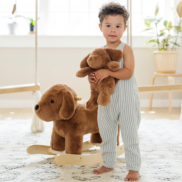 A smiling little boy toddler with curly hair holds a small plush dog while standing next to a larger plush dog rocker. Dressed in a striped jumpsuit, the child enjoys a playful moment in a bright and airy room filled with natural light. This heartwarming scene captures the joy of imaginative play, making it perfect for parents seeking cozy and engaging toys for their children.