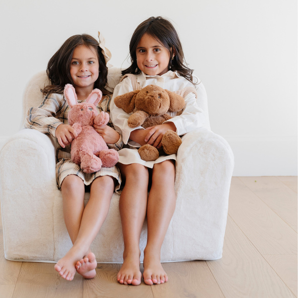 Two smiling girls sit side by side on a cozy white plush chair, each holding their stuffed animals—a pink bunny and a brown dog. Their joyful expressions capture the essence of childhood play and comfort, showcasing the perfect combination of stylish children's furniture and beloved plush toys. This delightful scene is ideal for parents searching for comfortable seating options and adorable toys for their children's play areas.