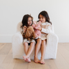 Two little girls sit side by side on a cozy white plush chair, each holding their stuffed animals—a pink bunny and a brown dog. Their joyful expressions capture the essence of childhood play and comfort, showcasing the perfect combination of stylish children's furniture and beloved plush toys. This delightful scene is ideal for parents searching for comfortable seating options and adorable toys for their children's play areas.