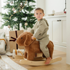 A little boy toddler in a cozy, olive-green outfit sits atop a plush brown dog rocker, gazing thoughtfully in a warmly decorated room with a Christmas tree. Gift boxes wrapped in festive paper are nestled nearby, creating a cheerful holiday atmosphere. This heartwarming scene highlights the joy of playtime and the perfect holiday gift for young children.