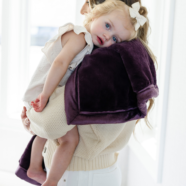 A mother holds her little girl who is laying on a Receiving Aubergine (deep purple) Colored Lush Saranoni Blanket. The soft blanket is a small blanket and a baby blanket or toddler blanket.