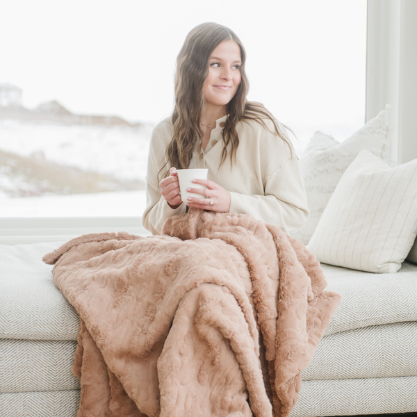 A woman smiles and sits with a rouge luxurious patterned faux fur throw blanket that is a Saranoni blanket. This ultra-soft, plush throw blanket adds a touch of elegance and warmth to any space, perfect as a soft throw blanket or fluffy blanket for lounging. Ideal for those seeking a high-quality artificial fur blanket or luxury blanket, this versatile and stylish soft blanket enhances both comfort and decor. Shop the best blankets and throws for ultimate relaxation and cozy vibes.