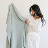 A woman holds up a Bamboni® blanket by Saranoni. This ultra-soft blanket is a sage colored blanket and made from a premium blend of bamboo rayon and poly microfiber, offering a lightweight, breathable, and fluffy feel. Perfect as a extra large blanket, luxury blanket, or soft blanket, it adds warmth and elegance to any living room, bedroom, or home décor setting. Designed for ultimate comfort, this buttery-soft Saranoni blanket is ideal for gifting. Enjoy this cozy blanket for years to come!