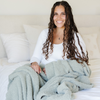 A woman sits with a Bamboni® blanket by Saranoni. This ultra-soft blanket is a sage colored blanket and made from a premium blend of bamboo rayon and poly microfiber, offering a lightweight, breathable, and fluffy feel. Perfect as a extra large blanket, luxury blanket, or soft blanket, it adds warmth and elegance to any living room, bedroom, or home décor setting. Designed for ultimate comfort, this buttery-soft Saranoni blanket is ideal for gifting. Enjoy this cozy blanket for years to come!