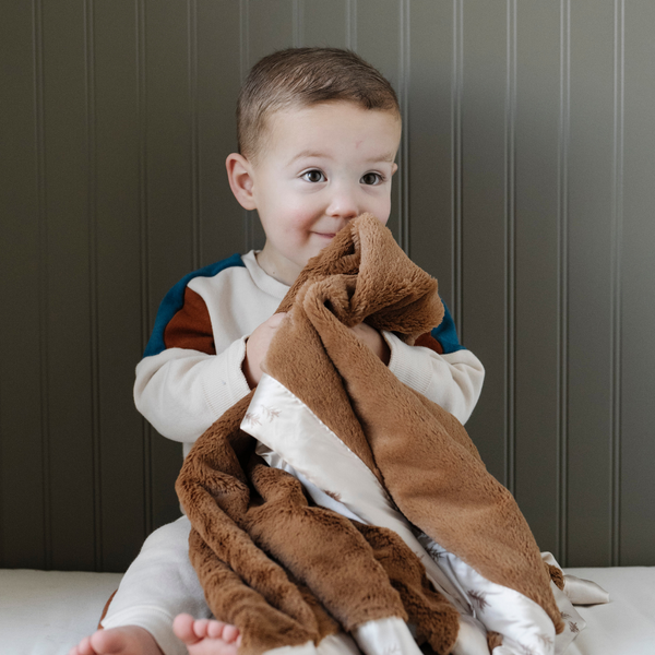 A little boy smiles and holds a satin back blanket. The lush blanket is brown with a cream satin border that has little trees on it. The satin back blanket is a lush blanket with a satin border and back. The soft blanket is a luxury blanket and a Saranoni blanket. This receiving blanket is an adorable baby blanket or toddler blanket and perfect for littles. 