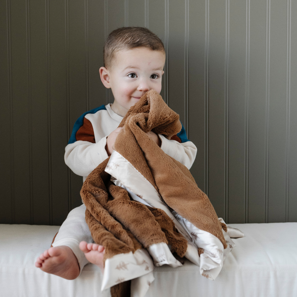 A little boy smiles and holds a satin back blanket. The lush blanket is brown with a cream satin border that has little trees on it. The satin back blanket is a lush blanket with a satin border and back. The soft blanket is a luxury blanket and a Saranoni blanket. This toddler blanket is an adorable baby blanket or toddler blanket and a great kids blanket.