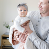 A father holds his daughter in a Saranoni blanket. The baby is wrapped in a beautifully soft and breathable muslin quilt, designed for both comfort and style. The quilt features a warm, earthy rust tone with delicate white botanical accents, adding a timeless and elegant touch. Its airy, multi-layered muslin fabric is crafted to become even softer with each wash, making it a perfect choice for snuggles, naps, and everyday moments. The soft blanket is a luxury blanket and a Saranoni blanket. 