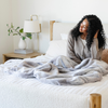 A woman sits on a bed with a Saranoni Grand Faux Fur blanket, featuring an elegant white and gray and brown tone and luxuriously soft texture. This cozy, throw blanket and faux fur blanket offers soothing weight, premium softness, timeless comfort, and is a soft blanket. Perfect as a cozy blanket and luxury blanket for snuggling or as a luxury bedroom accent, this Saranoni blanket elevates every cozy moment and is a cozy blanket.