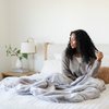 A woman sits with a Saranoni Grand Faux Fur blanket, featuring an elegant white and gray and brown tone and luxuriously soft texture. This cozy, throw blanket and faux fur blanket offers soothing weight, premium softness, timeless comfort, and is a soft blanket. Perfect as a cozy blanket and luxury blanket for snuggling or as a luxury bedroom accent, this Saranoni blanket elevates every cozy moment and is a cozy blanket.