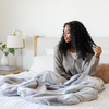 A woman sits with a Saranoni Grand Faux Fur blanket, featuring an elegant white and gray and brown tone and luxuriously soft texture. This cozy, extra large throw blanket and faux fur blanket offers soothing weight, premium softness, timeless comfort, and is a soft blanket. Perfect as a cozy blanket and luxury blanket for snuggling or as a luxury bedroom accent, this Saranoni blanket elevates every cozy moment and is a cozy blanket.