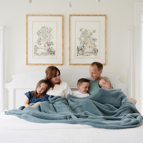 A husband, wife, and their kids sit under a Bamboni® blanket by Saranoni. This ultra-soft blanket is a light blue blanket and made from a premium blend of bamboo rayon and poly microfiber, offering a lightweight, breathable, and fluffy feel. Perfect as a extra large blanket, luxury blanket, or soft blanket, it adds warmth and elegance to any living room, bedroom, or home décor setting. Designed for ultimate comfort, this buttery-soft Saranoni blanket is ideal for gifting. Enjoy this cozy blanket!