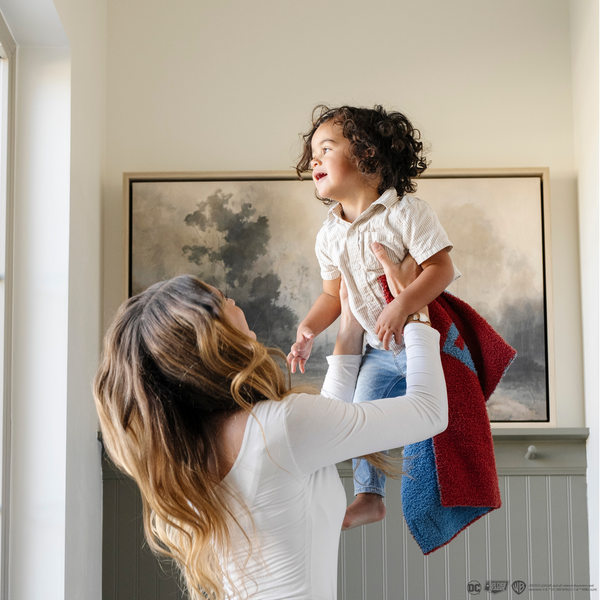 A mother plays with her little boy while holding a mini blanket with a Superman™ icon on it. The soft blanket is light blue and red. This kids blanket is a luxury blanket and a Saranoni blanket. The blanket is also a double-layer Bamboni® blanket. 