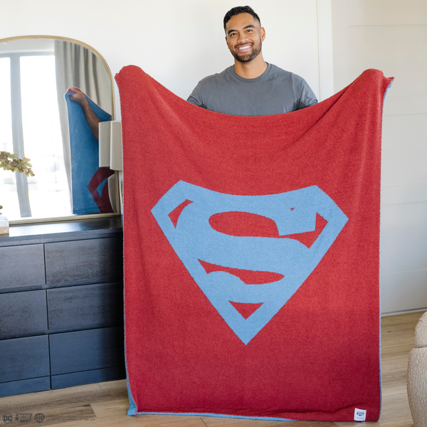 A man holds a blanket with a Superman™ icon on it. The soft blanket is light blue and red. This throw blanket is a luxury blanket and a Saranoni blanket. The blanket is also a double-layer Bamboni® blanket. 