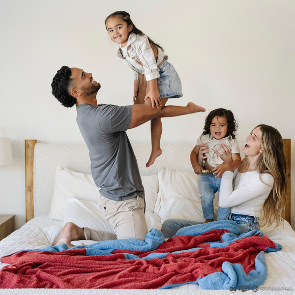 A husband, wife, and their little boy and little girl play on a bed. There is a blanket with a Superman™ icon on it by them. The soft blanket is light blue and red. This throw blanket is a luxury blanket and a Saranoni blanket. The blanket is also a double-layer Bamboni® blanket. 