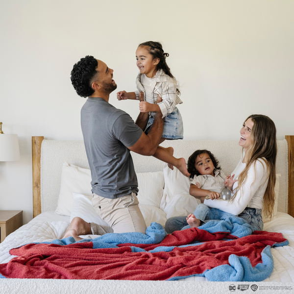 A husband, wife, and their little boy and little girl play on a bed. There is a blanket with a Superman™ icon on it by them. The soft blanket is light blue and red. This throw blanket is a luxury blanket and a Saranoni blanket. The blanket is also a double-layer Bamboni® blanket. 