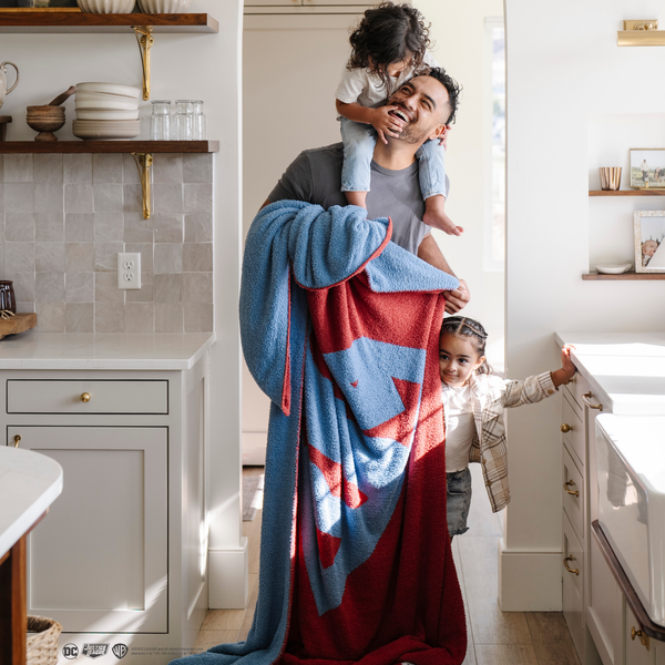 A man with his little boy and little girl holds a blanket with a Superman™ icon on it. The soft blanket is light blue and red. This throw blanket is a luxury blanket and a Saranoni blanket. The blanket is also a double-layer Bamboni® blanket. 