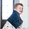 A little boy wrapped in a soft minky blanket showcasing Superman™ in action, complete with iconic red capes, the Superman™ emblem, comic-style speech bubbles, glasses, and stars. The white background enhances the vibrant red and blue superhero details, while the dark navy plush underside adds extra coziness. Ideal for snuggling, showcasing superhero pride, or adding a fun, adventurous touch to your decor. The soft blanket is a Saranoni blanket. 