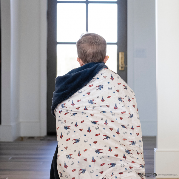 A little boy wrapped in a soft minky blanket showcasing Superman™ in action, complete with iconic red capes, the Superman™ emblem, comic-style speech bubbles, glasses, and stars. The white background enhances the vibrant red and blue superhero details, while the dark navy plush underside adds extra coziness. Ideal for snuggling, showcasing superhero pride, or adding a fun, adventurous touch to your decor. The soft blanket is a Saranoni blanket. 