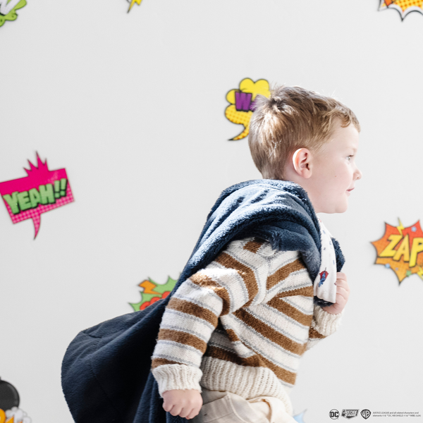 A little boy runs with a soft minky blanket showcasing Superman™ in action, complete with iconic red capes, the Superman™ emblem, comic-style speech bubbles, glasses, and stars. The white background enhances the vibrant red and blue superhero details, while the dark navy plush underside adds extra coziness. Ideal for snuggling, showcasing superhero pride, or adding a fun, adventurous touch to your decor. The soft blanket is a Saranoni blanket. 