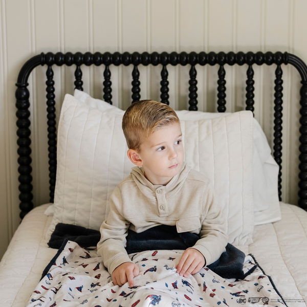 A little boy sits on a bed with a soft minky blanket showcasing Superman™ in action, complete with iconic red capes, the Superman™ emblem, comic-style speech bubbles, glasses, and stars. The white background enhances the vibrant red and blue superhero details, while the dark navy plush underside adds extra coziness. Ideal for snuggling, showcasing superhero pride, or adding a fun, adventurous touch to your decor. The soft blanket is a Saranoni blanket. 