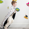 A little boy runs with a soft minky blanket showcasing Superman™ in action, complete with iconic red capes, the Superman™ emblem, comic-style speech bubbles, glasses, and stars. The white background enhances the vibrant red and blue superhero details, while the dark navy plush underside adds extra coziness. Ideal for snuggling, showcasing superhero pride, or adding a fun, adventurous touch to your decor. The soft blanket is a Saranoni blanket. 