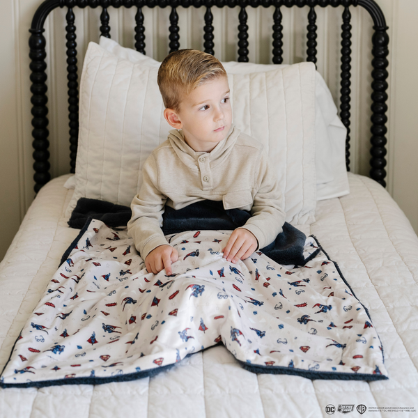 A little boy on a bed with a soft minky blanket showcasing Superman™ in action, complete with iconic red capes, the Superman™ emblem, comic-style speech bubbles, glasses, and stars. The white background enhances the vibrant red and blue superhero details, while the dark navy plush underside adds extra coziness. Ideal for snuggling, showcasing superhero pride, or adding a fun, adventurous touch to your decor. The soft blanket is a Saranoni blanket. 