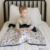 A little boy on a bed with a soft minky blanket showcasing Superman™ in action, complete with iconic red capes, the Superman™ emblem, comic-style speech bubbles, glasses, and stars. The white background enhances the vibrant red and blue superhero details, while the dark navy plush underside adds extra coziness. Ideal for snuggling, showcasing superhero pride, or adding a fun, adventurous touch to your decor. The soft blanket is a Saranoni blanket. 