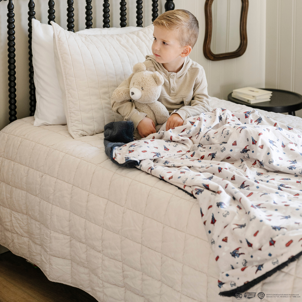 A little boy on his bed with a bear stuffed animal and a soft minky blanket showcasing Superman™ in action, complete with iconic red capes, the Superman™ emblem, comic-style speech bubbles, glasses, and stars. The white background enhances the vibrant red and blue superhero details, while the dark navy plush underside adds extra coziness. Ideal for snuggling, showcasing superhero pride, or adding a fun, adventurous touch to your decor. The soft blanket is a Saranoni blanket. 