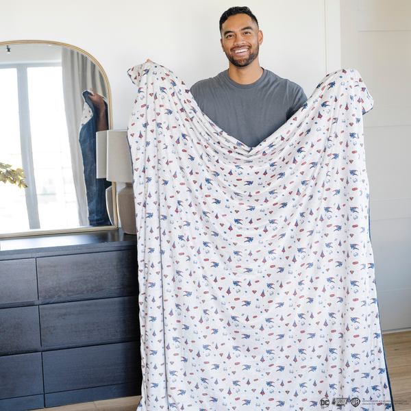 A man holds a soft minky blanket showcasing Superman™ in action, complete with iconic red capes, the Superman™ emblem, comic-style speech bubbles, glasses, and stars. The white background enhances the vibrant red and blue superhero details, while the dark navy plush underside adds extra coziness. Ideal for snuggling, showcasing superhero pride, or adding a fun, adventurous touch to your decor. The soft blanket is a Saranoni blanket. 