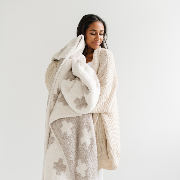 A woman smiles and holds a luxurious double-layer Bamboni® blanket by Saranoni, featuring a cream and taupe blanket with a swiss cross pattern. This ultra-soft blanket is crafted from bamboo rayon and poly microfiber, making it lightweight, breathable, and fluffy. Ideal as a cozy throw blanket, luxury blanket, plush throw, soft blanket, or stylish home accent, this Saranoni blanket brings warmth, comfort, and timeless elegance to any living room or bedroom.