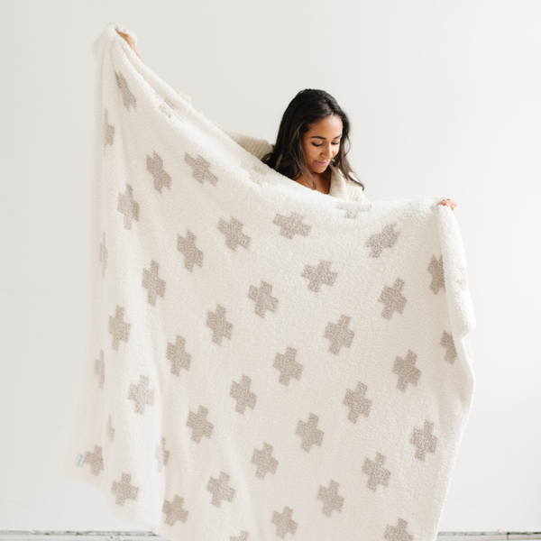 A woman holds up a luxurious double-layer Bamboni® blanket by Saranoni, featuring a cream and taupe blanket with a swiss cross pattern. This ultra-soft blanket is crafted from bamboo rayon and poly microfiber, making it lightweight, breathable, and fluffy. Ideal as a cozy throw blanket, luxury blanket, plush throw, soft blanket, or stylish home accent, this Saranoni blanket brings warmth, comfort, and timeless elegance to any living room or bedroom.