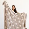 A smiling woman holds a luxurious double-layer Bamboni® blanket by Saranoni, featuring a cream and taupe blanket with a swiss cross pattern. This ultra-soft blanket is crafted from bamboo rayon and poly microfiber, making it lightweight, breathable, and fluffy. Ideal as a cozy blanket, extra large blanket, luxury blanket, soft blanket, or stylish home accent, this Saranoni blanket brings warmth, comfort, and timeless elegance to any living room or bedroom.