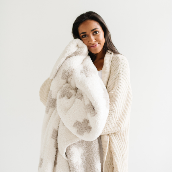 A woman smiles and holds a luxurious double-layer Bamboni® blanket by Saranoni, featuring a cream and taupe blanket with a swiss cross pattern. This ultra-soft blanket is crafted from bamboo rayon and poly microfiber, making it lightweight, breathable, and fluffy. Ideal as a cozy throw blanket, luxury blanket, plush throw, soft blanket, or stylish home accent, this Saranoni blanket brings warmth, comfort, and timeless elegance to any living room or bedroom.