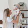 A mother holds up her happy baby girl and a double-layer Bamboni® baby blanket featuring a cream and taupe blanket with a swiss cross pattern. Made from Saranoni’s signature stretchy, cloud-like Bamboni® fabric, this luxury baby blanket provides comfort for naps, tummy time, and on-the-go snuggles. The soft blanket is a receiving blanket and Saranoni blanket. 