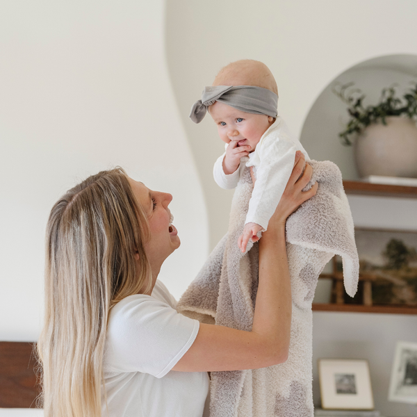 A mother smiles and holds up her happy baby girl and a double-layer Bamboni® baby blanket featuring a cream and taupe blanket with a swiss cross pattern. Made from Saranoni’s signature stretchy, cloud-like Bamboni® fabric, this luxury baby blanket provides comfort for naps, tummy time, and on-the-go snuggles. The soft blanket is a receiving blanket and Saranoni blanket. 