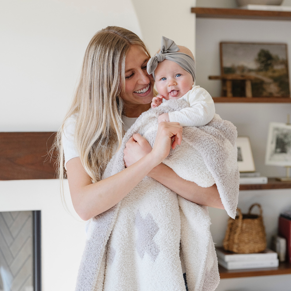 A mother smiles and holds her happy baby girl and a double-layer Bamboni® baby blanket featuring a cream and taupe blanket with a swiss cross pattern. Made from Saranoni’s signature stretchy, cloud-like Bamboni® fabric, this luxury baby blanket provides comfort for naps, tummy time, and on-the-go snuggles. The soft blanket is a receiving blanket and Saranoni blanket. 