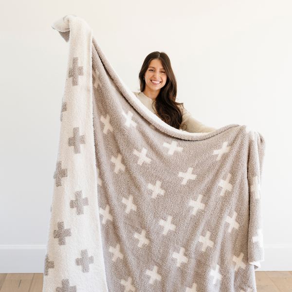 A smiling woman holds up a luxurious double-layer Bamboni® blanket by Saranoni, featuring a cream and taupe blanket with a swiss cross pattern. This ultra-soft blanket is crafted from bamboo rayon and poly microfiber, making it lightweight, breathable, and fluffy. Ideal as a cozy blanket, extra large blanket, luxury blanket, soft blanket, or stylish home accent, this Saranoni blanket brings warmth, comfort, and timeless elegance to any living room or bedroom.