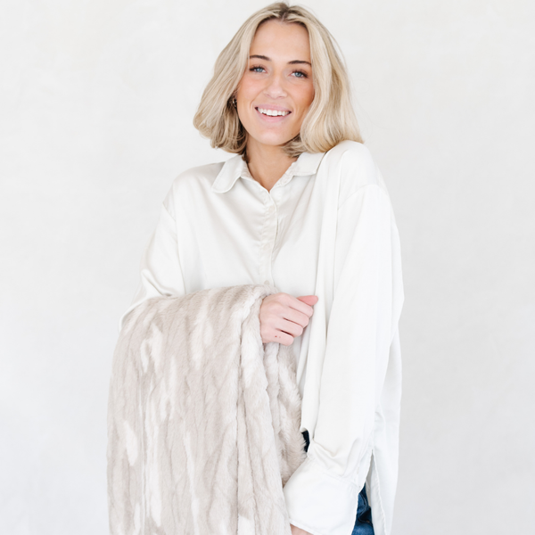 A woman smiles while holding a Saranoni blanket, specifically the tan marbled faux fur/minky throw, highlighting its ultra-soft texture and luxurious design. This plush throw blanket features a dual-texture blend of silky minky and cozy faux fur, creating the ultimate cozy throw blanket for home decor. Perfect luxury blanket for lounging or adding style to any space. Soft blanket, cozy blanket, plush throw—experience unmatched warmth with a Saranoni throw blanket.