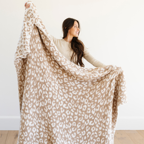 A woman holds up a luxurious double-layer Bamboni® blanket by Saranoni, featuring tan leopard print that is white and tan. This ultra-soft blanket is crafted from bamboo rayon and poly microfiber, making it lightweight, breathable, and fluffy. Ideal as a cozy blanket, extra large blanket, luxury blanket, soft blanket, or stylish home accent, this Saranoni blanket brings warmth, comfort, and timeless elegance to any living room or bedroom.