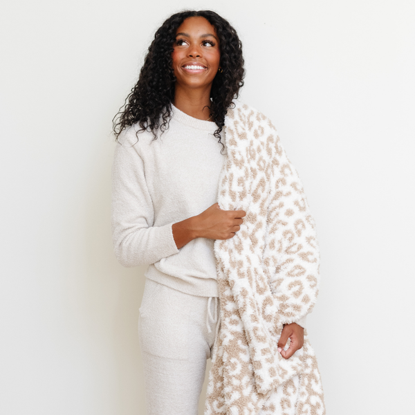 A woman smiles and holds a luxurious double-layer Bamboni® blanket by Saranoni, featuring tan leopard print that is white and tan. This ultra-soft blanket is crafted from bamboo rayon and poly microfiber, making it lightweight, breathable, and fluffy. Ideal as a cozy throw blanket, luxury blanket, plush throw, soft blanket, or stylish home accent, this Saranoni blanket brings warmth, comfort, and timeless elegance to any living room or bedroom.