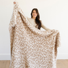 A woman smiles and holds up a luxurious double-layer Bamboni® blanket by Saranoni, featuring tan leopard print that is white and tan. This ultra-soft blanket is crafted from bamboo rayon and poly microfiber, making it lightweight, breathable, and fluffy. Ideal as a cozy blanket, extra large blanket, luxury blanket, soft blanket, or stylish home accent, this Saranoni blanket brings warmth, comfort, and timeless elegance to any living room or bedroom.