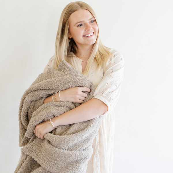 A woman smiles and holds a Bamboni® blanket by Saranoni. This ultra-soft blanket is a taupe blanket and made from a premium blend of bamboo rayon and poly microfiber, offering a lightweight, breathable, and fluffy feel. Perfect as a throw blanket, luxury blanket, plush throw, or soft blanket, it adds warmth and elegance to any living room, bedroom, or home décor setting. Designed for ultimate comfort, this buttery-soft Saranoni blanket is ideal for gifting. Enjoy this cozy blanket for years to come!