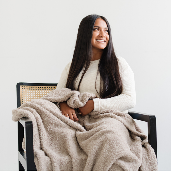 A woman smiles and sits with a Bamboni® blanket by Saranoni. This ultra-soft blanket is a taupe blanket and made from a premium blend of bamboo rayon and poly microfiber, offering a lightweight, breathable, and fluffy feel. Perfect as a extra large blanket, luxury blanket, or soft blanket, it adds warmth and elegance to any living room, bedroom, or home décor setting. Designed for ultimate comfort, this buttery-soft Saranoni blanket is ideal for gifting. Enjoy this cozy blanket for years to come! 